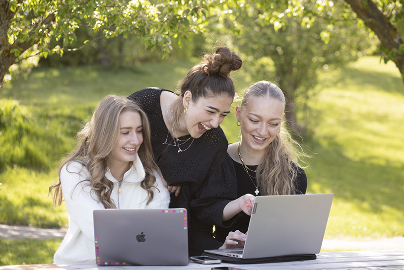 Tre elever fra en af NEG's afdelinger sidder udenfor og kigger på en computer. Pigerne ser meget glade ud. 