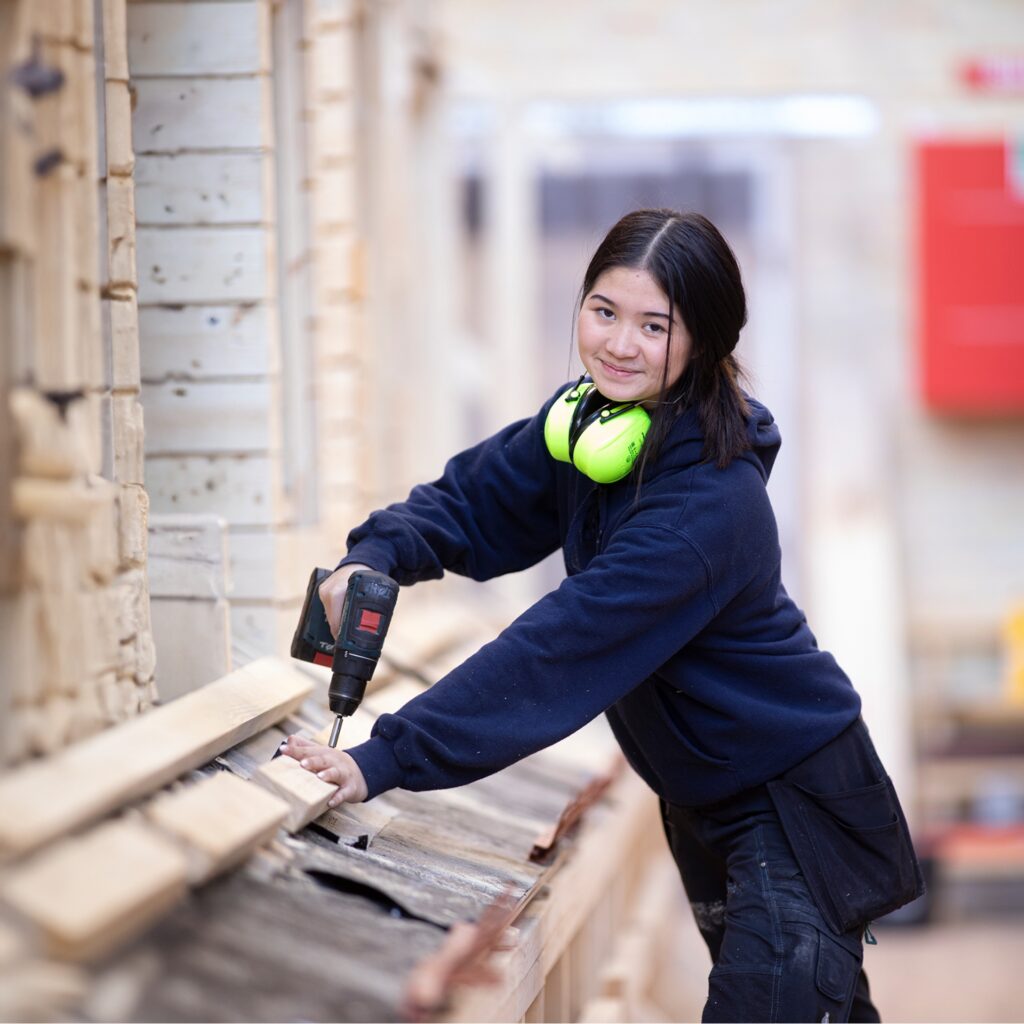 En elev fra tømreruddannelsen på NEG står og arbejder på et projekt. Hun har høreværn om halsen og står med en boremaskine i hånden.
