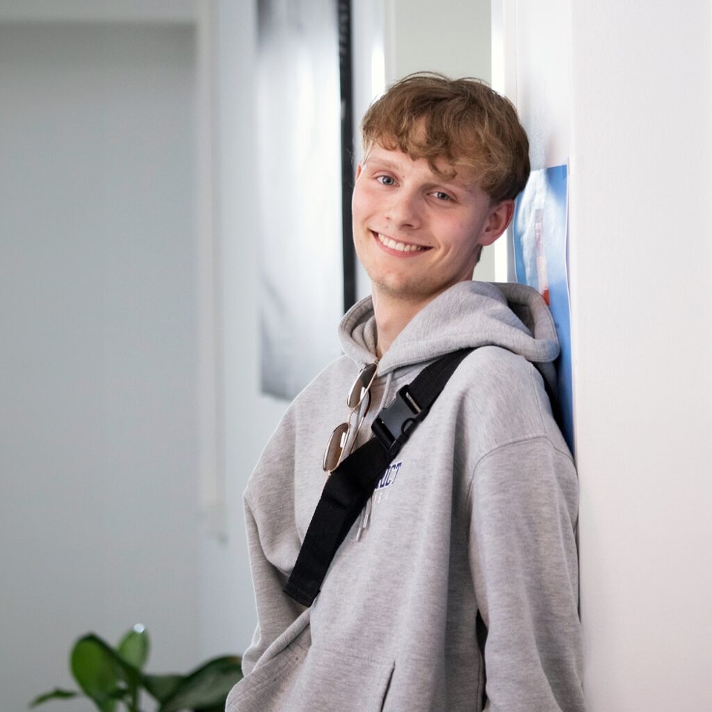 Et portræt af en elev fra NEG. Drengen står og læner ryggen op af en væg. Han er iført en sweatshirt og har en taskerem hængende over brystet.