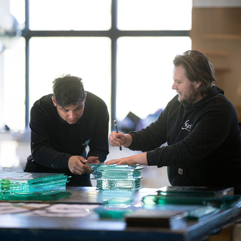 To elever fra glarmester uddannelsen står og arbejder med nogle glasplader.