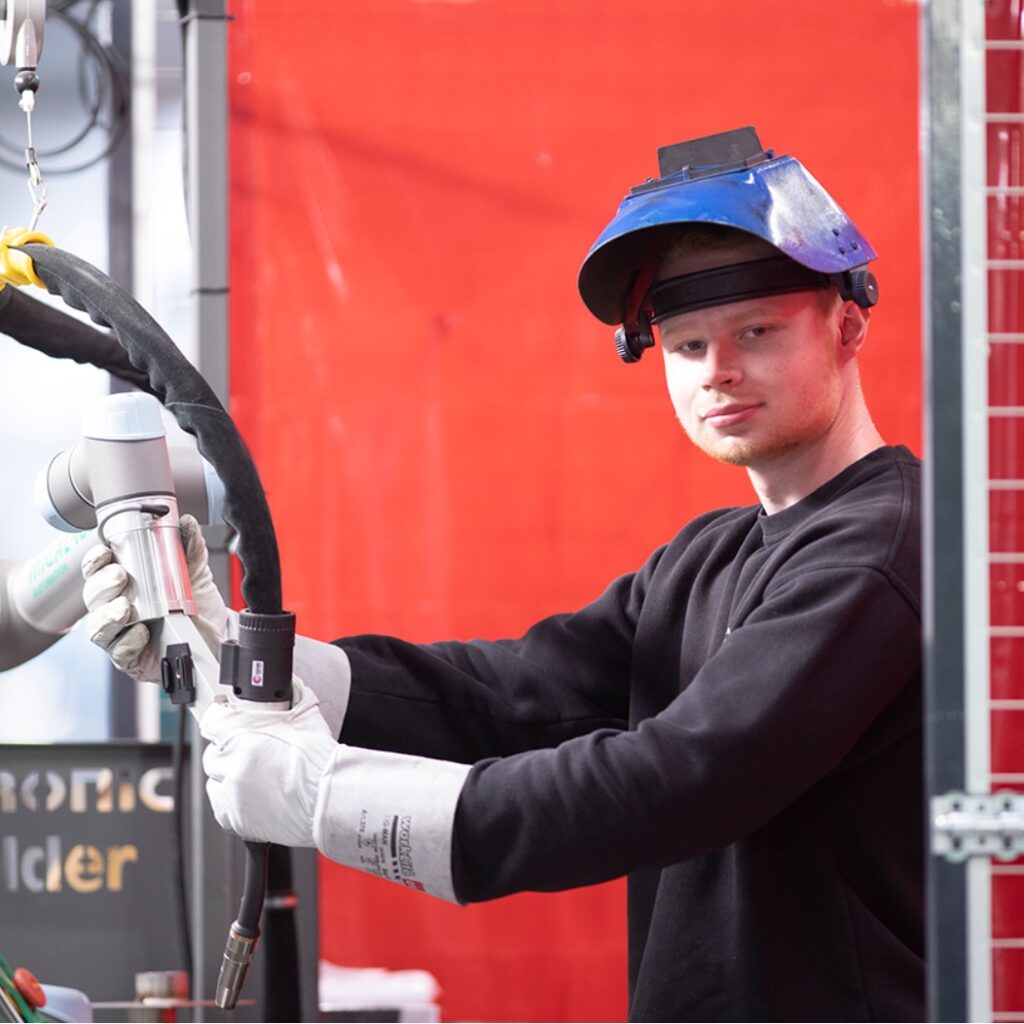En elev fra NEG er iført arbejdstøj, hjelm og handsker. Han står og arbejder med en maskine.